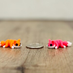 May include: Two small fox figurines, one orange and one pink, each featuring white paws and a white-tipped tail, positioned on either side of a silver-coloured coin for scale. The foxes have black eyes and are in a lying down position.