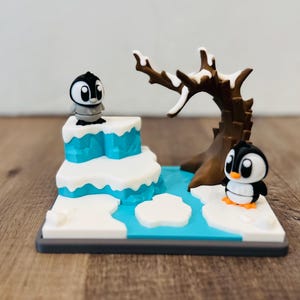 May include: A miniature diorama featuring two penguin figurines in a snowy landscape. The scene includes a blue water feature, white snow, and a brown tree. One penguin stands atop a snowy ledge, while the other stands on the ground.