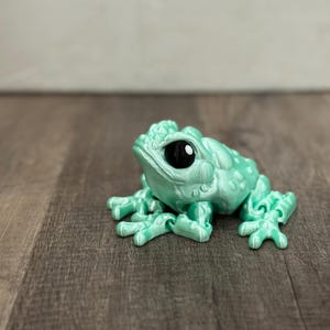 May include: A mint green frog figurine with large black eyes. The frog has a textured skin and is posed with its legs spread out. The figurine is sitting on a wooden surface.