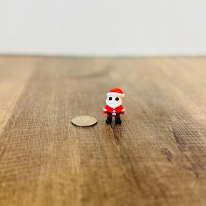 Pode incluir: Uma figura em miniatura do Pai Natal, vestindo um traje vermelho, botas pretas e um chapéu com acabamento branco. Uma moeda está ao lado para referência de tamanho. O fundo é uma superfície de madeira.