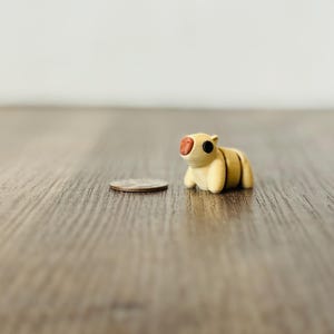 May include: A small, handcrafted, light yellow figurine of an animal with a red nose and black eyes. The figurine is next to a quarter coin on a wooden surface. The animal has a segmented body.