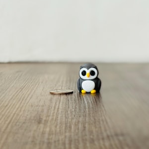 May include: A small, black and gray owl figurine with large white eyes and yellow feet. A coin is next to the owl for size comparison. The figurine is on a wooden surface.
