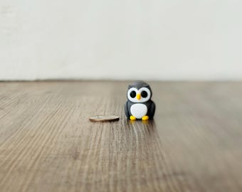 May include: A small, black and gray owl figurine with large white eyes and yellow feet. A coin is next to the owl for size comparison. The figurine is on a wooden surface.