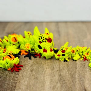 May include: A neon green articulated dragon toy with black, orange, and red accents. The dragon has a detailed head and body, with multiple segments. The toy is posed on a wooden surface.