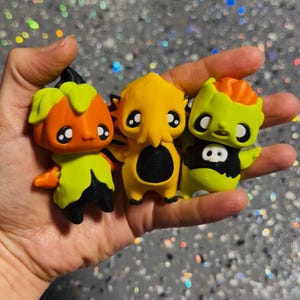 May include: Three colorful, cartoonish figurines are held in a hand. The figurines are a pumpkin, a yellow creature, and a zombie. The pumpkin is orange and green, the yellow creature has black accents, and the zombie is green and black.