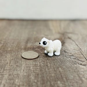 May include: A small, white polar bear figurine with black eyes and paws stands next to a quarter on a wooden surface. The figurine is detailed and appears to be made of plastic. The background is a blurred wooden texture.