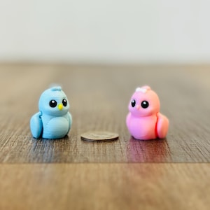 May include: Two small, colorful bird figurines. One is light blue with a yellow beak, the other is pink. They are next to a coin on a wooden surface. The birds have large black eyes.
