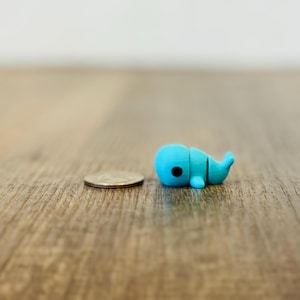 May include: A small, light blue whale figurine with a black eye, lying on a wooden surface next to a quarter. The whale is approximately 1 inch long, with a segmented design. The background is a blurred, neutral color.