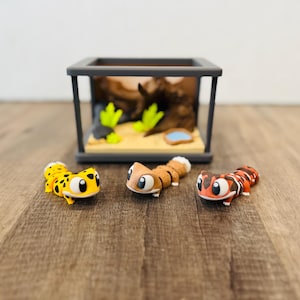 以下が含まれることがあります： 大きな目をした3匹のカラフルなヤモリのフィギュアが、ミニチュアテラリウムの前に展示されています。ヤモリは、黒い斑点のある黄色、タンストライプのある茶色、黒いストライプのある赤色です。テラリウムは、詳細なインテリアを備えたグレーです。