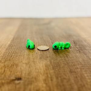 May include: Two small, bright green chameleon figurines with black eyes are displayed on a wooden surface. A coin is placed between the two figurines. The chameleon on the right is segmented, and the other is in a curled position.