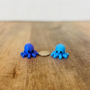 May include: Two small, blue octopus figurines with black eyes and smiling faces sit on a wooden surface. One is a darker blue, the other a lighter blue. A coin is between them.