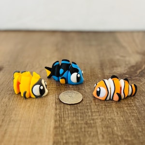 May include: Three colorful, handmade clay fish figurines. One is yellow and black, another is blue and black, and the third is orange, white, and black. A coin is placed between the figurines on a wooden surface.