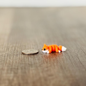 May include: A miniature orange fox figurine with white accents, lying next to a quarter coin. The fox has black eyes and is made of a smooth material. The figurine is approximately 1 inch long.