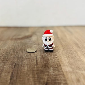 May include: A miniature Santa Claus figurine with a red hat and suit, white beard, and black boots. The figure is standing next to a coin on a wooden surface. A festive Christmas decoration.