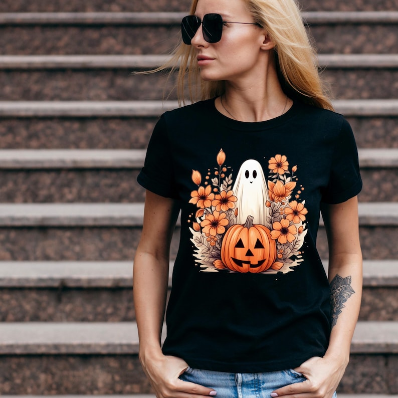 May include: Black t-shirt with a white ghost and orange pumpkin in a floral arrangement. The ghost is standing behind the pumpkin, which has a carved jack-o'-lantern face.