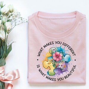 Op de afbeelding: Roze T-shirt met een kleurrijk puzzelstuk ontwerp en de tekst "Wat je anders maakt, maakt je mooi."