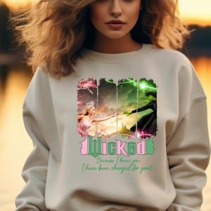 May include: A white sweatshirt featuring a graphic print of two women from the musical Wicked. The text "Wicked" is in pink and green lettering. The text "Because I knew you I have been changed for good" is in green lettering.