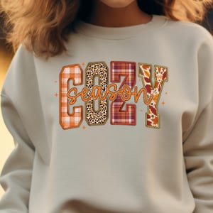 Puede incluir: Sudadera gris claro con las palabras "COZY SEASON" en un dise&ntilde;o con tem&aacute;tica oto&ntilde;al. Las letras est&aacute;n rellenas de cuadros, estampado de leopardo y patrones de hojas de oto&ntilde;o. La sudadera tiene cuello redondo.
