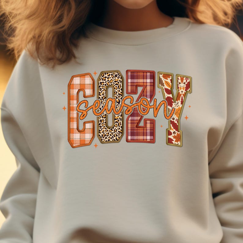 Puede incluir: Sudadera gris claro con las palabras "COZY SEASON" en un dise&ntilde;o con tem&aacute;tica oto&ntilde;al. Las letras est&aacute;n rellenas de cuadros, estampado de leopardo y patrones de hojas de oto&ntilde;o. La sudadera tiene cuello redondo.