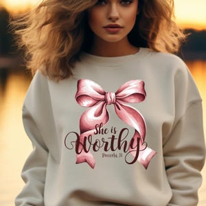 May include: A light beige sweatshirt with a pink bow and the text "She is worthy Proverbs 31" printed on the front.