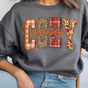 Puede incluir: Una sudadera gris con la frase "COZY SEASON". Las letras est&aacute;n decoradas con motivos oto&ntilde;ales, incluyendo cuadros escoceses, estampado de leopardo y hojas. La sudadera se combina con unos vaqueros azules.
