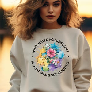 Op de afbeelding: Een lichtbeige sweatshirt met een kleurrijk puzzelstukontwerp en de tekst "Wat je anders maakt, maakt je mooi."