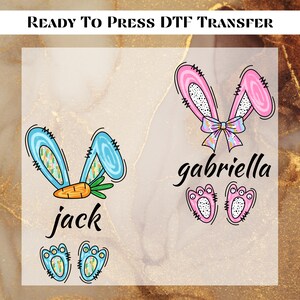 May include: Two iron-on transfers featuring bunny ears and paw prints. The transfer on the left is blue and features a carrot and the name "jack". The transfer on the right is pink and features a bow and the name "gabriella".