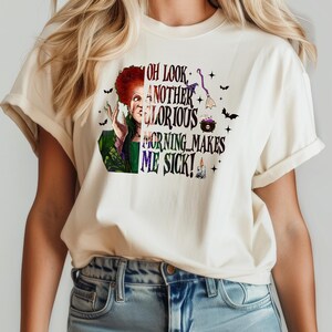May include: Cream-colored t-shirt featuring a graphic of a character with red hair and green clothing. The text on the shirt reads "OH LOOK, ANOTHER GLORIOUS MORNING MAKES ME SICK!" with additional Halloween-themed illustrations.
