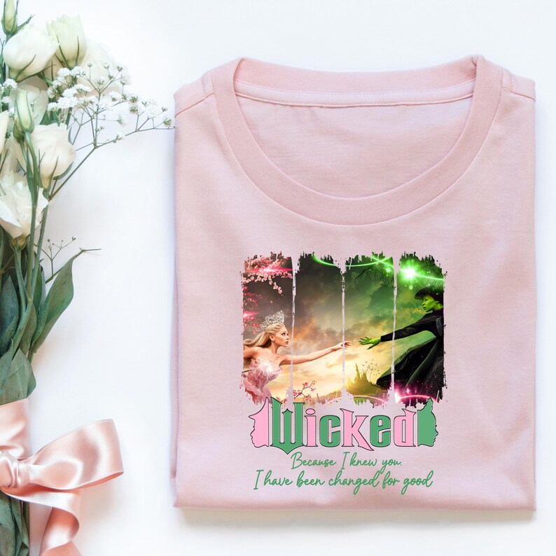 May include: Pink t-shirt featuring a graphic print of two women from the musical Wicked. The text "Wicked" is in green and pink, and the text "Because I knew you I have been changed for good" is in black.