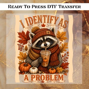 May include: A DTF transfer featuring a raccoon wearing a knit hat and scarf, holding a coffee cup with a pumpkin design. The text reads "I IDENTIFY AS A PROBLEM" and "READY TO PRESS DTF TRANSFER". Surrounded by autumn leaves, berries, and pumpkins.
