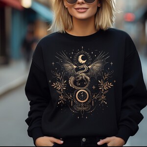 May include: Black sweatshirt with a gold graphic of a dragon with wings spread, a crescent moon, and stars on a dark background.
