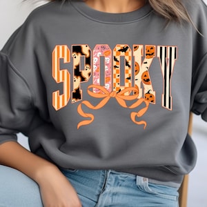 May include: Gray sweatshirt with the word "SPOOKY" in orange, black, and white block letters. The letters are decorated with Halloween-themed patterns, including ghosts, bats, and pumpkins. An orange bow is below the word.