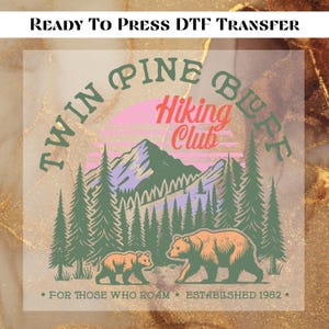 May include: A DTF transfer featuring a nature scene with the text "Twin Pine Buff Hiking Club." The design includes a mountain range, pine trees, and two bears. The text "For those who roam" and "Established 1982" are also included.