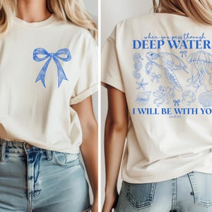 Peut inclure: T-shirt crème avec un motif de nœud bleu sur le devant. Le dos du t-shirt présente un texte bleu qui dit "When you pass through deep water, I will be with you." avec des illustrations de la vie marine.