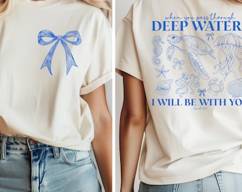 May include: Cream-colored t-shirt with a blue bow graphic on the front. The back of the shirt features blue text that reads "When you pass through deep water, I will be with you." with sea life illustrations.