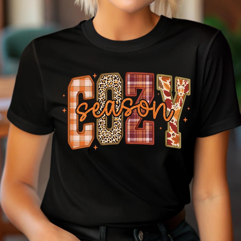 Puede incluir: Camiseta negra con las palabras "COZY SEASON" en un dise&ntilde;o con tem&aacute;tica oto&ntilde;al. Las letras est&aacute;n rellenas de cuadros, estampado de leopardo y hojas de oto&ntilde;o. La palabra "season" est&aacute; en cursiva, con peque&ntilde;os acentos de estrellas.