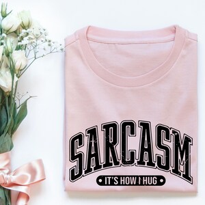 May include: A pink t-shirt with black text that reads "SARCASM IT'S HOW I HUG".