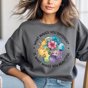Op de afbeelding: Grijs sweatshirt met een kleurrijk puzzelstuk ontwerp en de tekst "Wat je anders maakt, maakt je mooi."