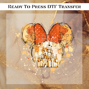 May include: A Ready To Press DTF Transfer featuring a sketch-style illustration of a castle beneath a large, orange, polka-dotted bow with mouse ears. The design includes yellow and orange paint splatters and ink-like outlines on a gold background.