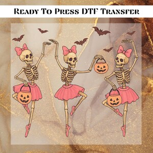 May include: Ready to Press DTF Transfer. A Halloween-themed design featuring three skeleton ballerinas in pink tutus and bows, holding jack-o'-lantern buckets. Bats are scattered across the background. The design is on a gold and white marbled background.