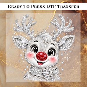 May include: A Ready To Press DTF Transfer featuring a cartoon reindeer with a bright red nose, large dark eyes, and pink cheeks. The reindeer is covered in a silver rhinestone-like texture, including its antlers and a scarf with a flower pin.