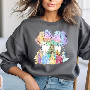 May include: A gray sweatshirt with a colorful graphic of Disney princesses in front of Cinderella's castle. The princesses are wearing their signature dresses and the graphic is surrounded by a pink and white bow with polka dots.