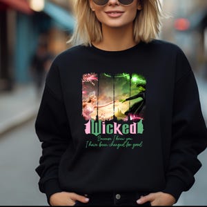 May include: Black sweatshirt featuring a graphic print of the Wicked musical characters Elphaba and Glinda. The text "Wicked" is in pink and green lettering. The text "Because I knew you, I have been changed for good" is printed below the graphic.