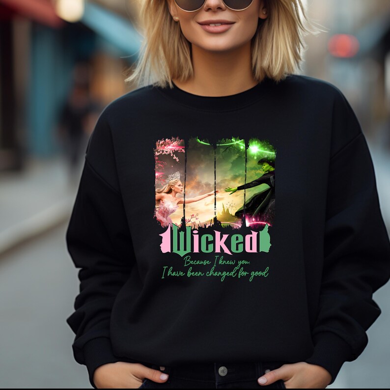 May include: Black sweatshirt featuring a graphic print of the Wicked musical characters Elphaba and Glinda. The text "Wicked" is in pink and green lettering. The text "Because I knew you, I have been changed for good" is printed below the graphic.