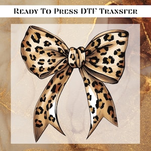 May include: A ready-to-press DTF transfer featuring a large leopard print bow. The bow is brown and black with a white background.