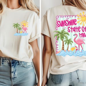May include: Cream-colored t-shirt featuring a tropical design. The front displays a flamingo, palm trees, and a sun graphic. The back of the shirt has the text "Sunshine State Of Mind" with a flamingo, sun, and palm trees. The shirt is paired with blue jeans.