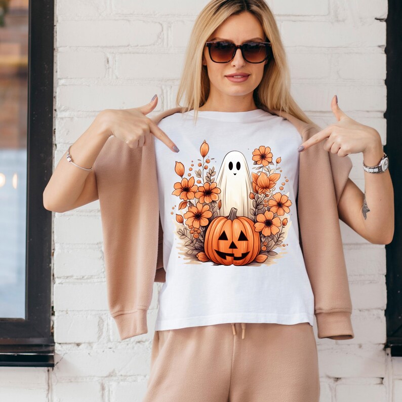 May include: White t-shirt with a graphic of a ghost standing on top of a jack-o'-lantern pumpkin surrounded by orange flowers.