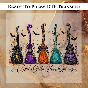 May include: A Ready To Press DTF Transfer featuring five stylized witch brooms in black, purple, orange, and teal, decorated with stars, bows, bats, and spiderwebs. The text A Girl's Gotta Have Options is written in a cursive script at the bottom.