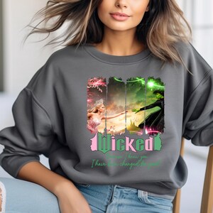 May include: A grey sweatshirt featuring a graphic print of the Wicked musical characters Elphaba and Glinda. The text "Wicked" is in green and pink lettering. The text "Because I knew you, I have been changed for good" is in pink lettering.