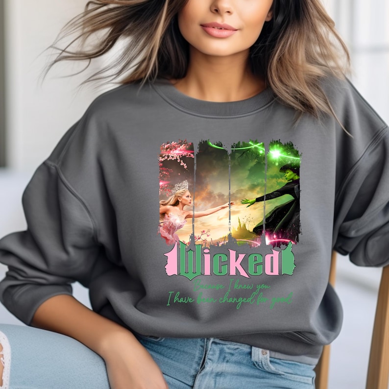 May include: A grey sweatshirt featuring a graphic print of the Wicked musical characters Elphaba and Glinda. The text "Wicked" is in green and pink lettering. The text "Because I knew you, I have been changed for good" is in pink lettering.